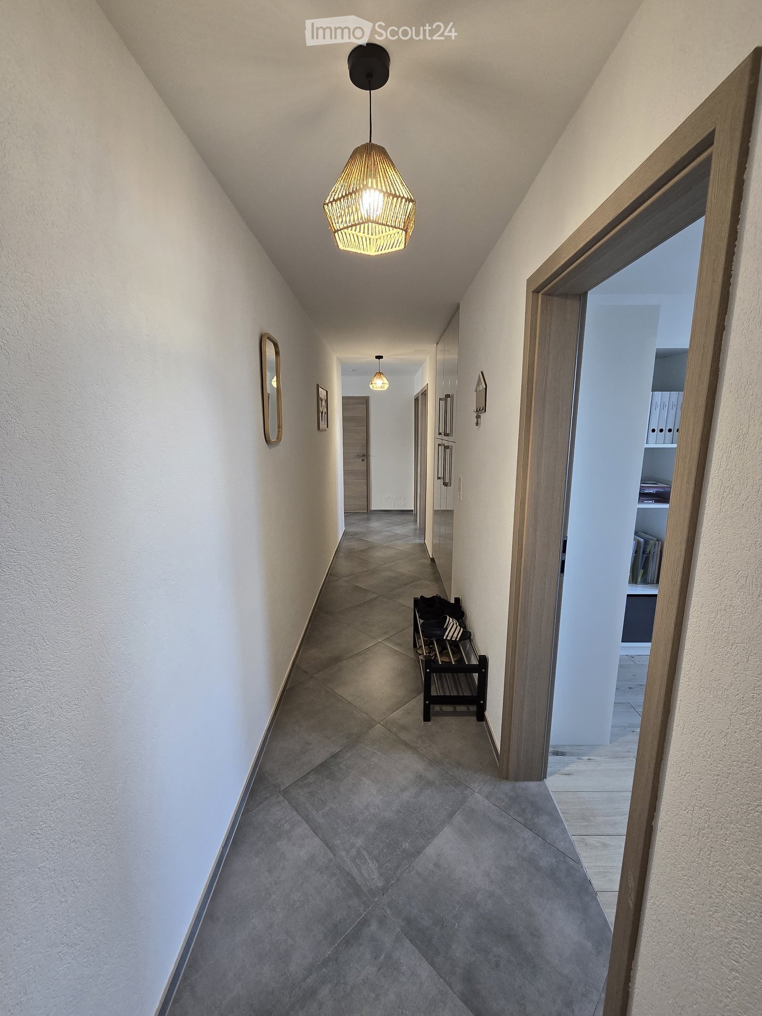 A modern hallway with tiled flooring, white walls, and a hanging woven pendant light fixture. A small bench with folded items sits against the wall, and a partially open door reveals a glimpse of a room with shelves and books.