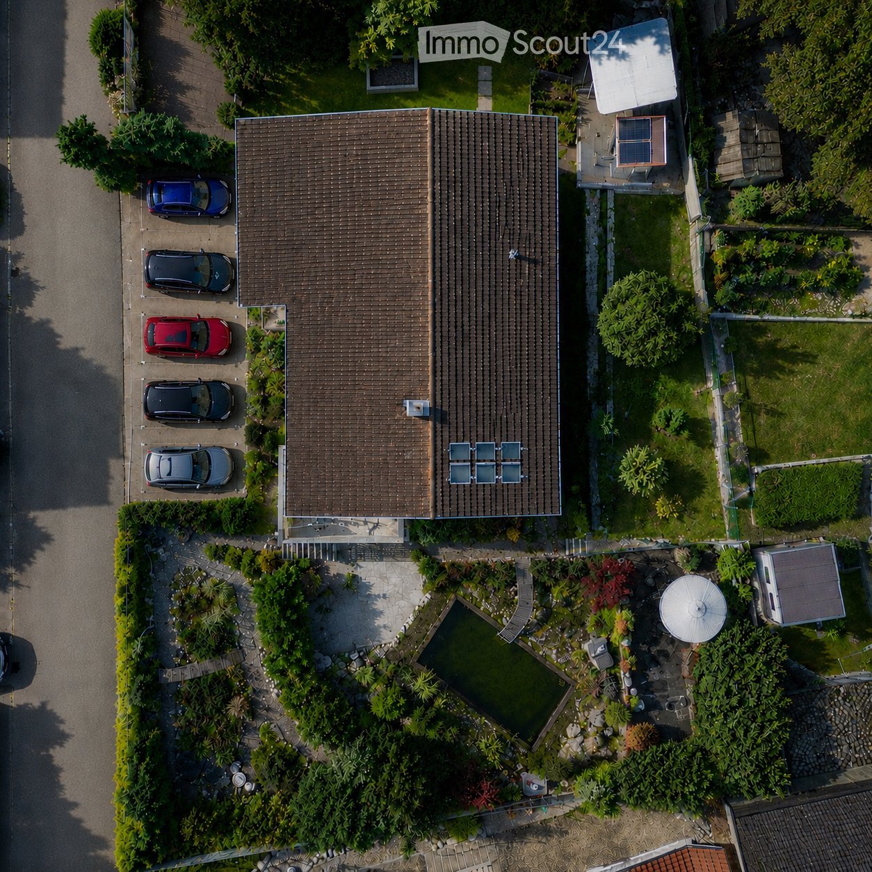 Aerial view of a two-story house with a brown tiled roof, surrounded by a well-maintained garden, a swimming pool, and a parking area with several cars.