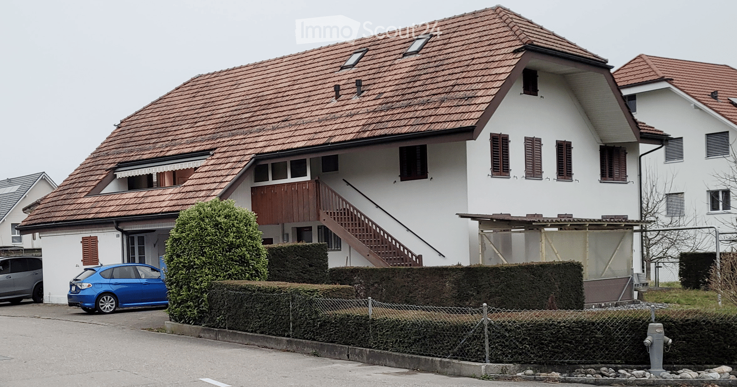 Two-story house with a red-tiled roof, white exterior, and multiple windows with shutters. A wooden staircase leads to an upper-level balcony. A blue car is parked in front, and the house is surrounded by neatly trimmed hedges and a chain-link fence.