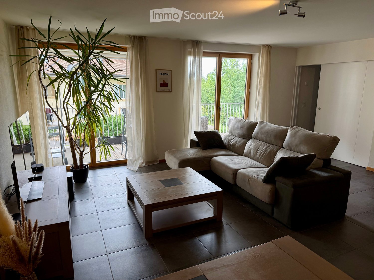 Modern living room with a large sectional sofa, wooden coffee table, TV stand, laptop, and a tall potted plant near the sliding glass doors leading to a balcony.