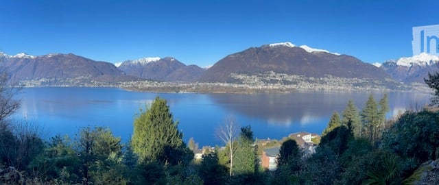 Panoramic view of a lake surrounded by snow-capped mountains, with trees and a small settlement visible on the shore.
