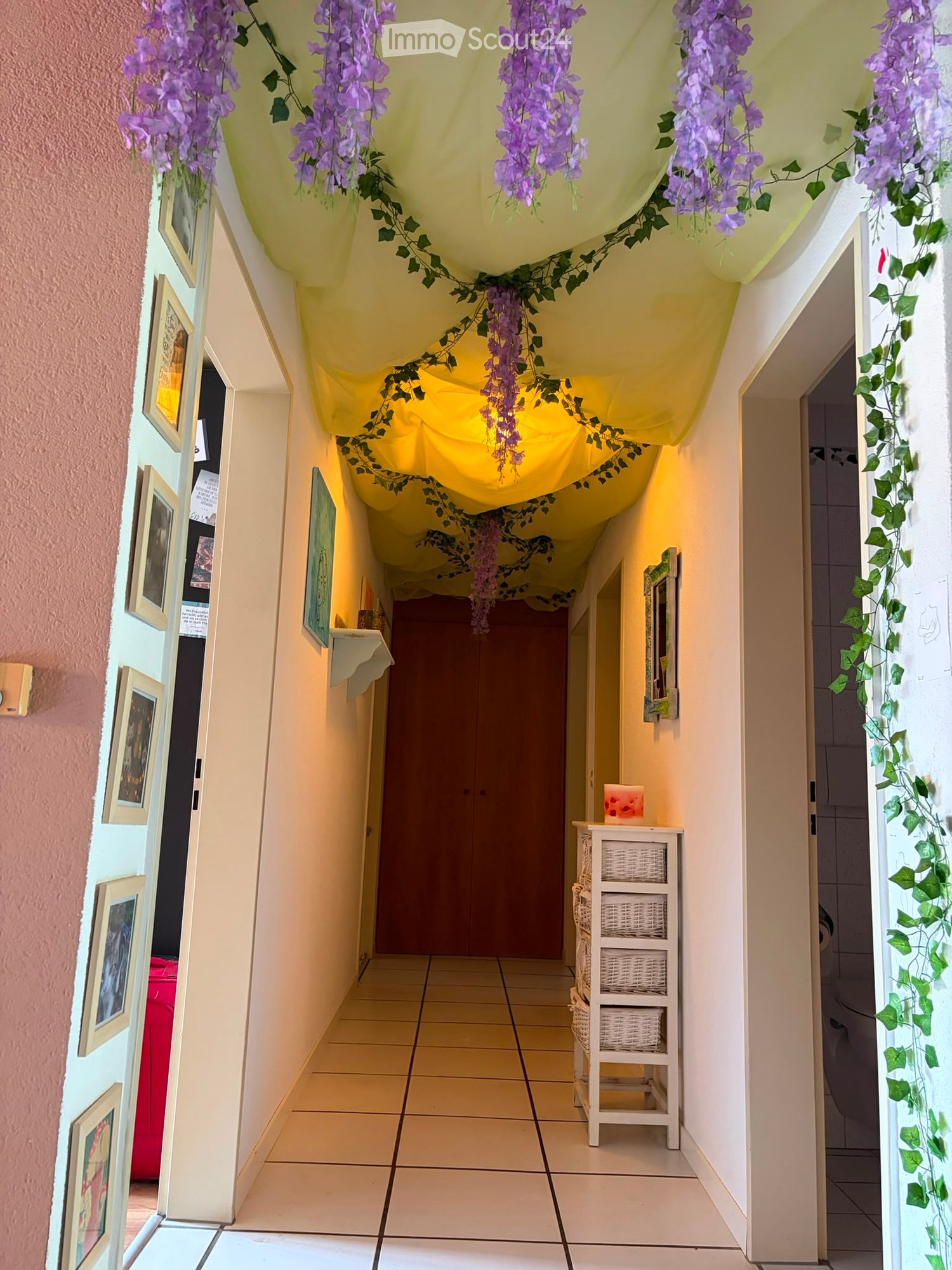 A narrow hallway with tiled flooring, light-colored walls, and a yellow draped ceiling adorned with hanging purple flowers. Several framed pictures are displayed on the left wall, and a small white shelf with wicker baskets and a candle is positioned on t