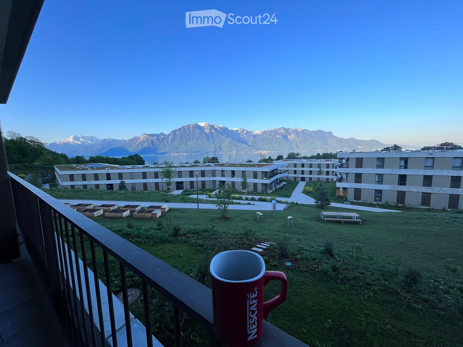 Balcony overlooking a grassy area with raised garden beds, benches, and distant mountains and lake.