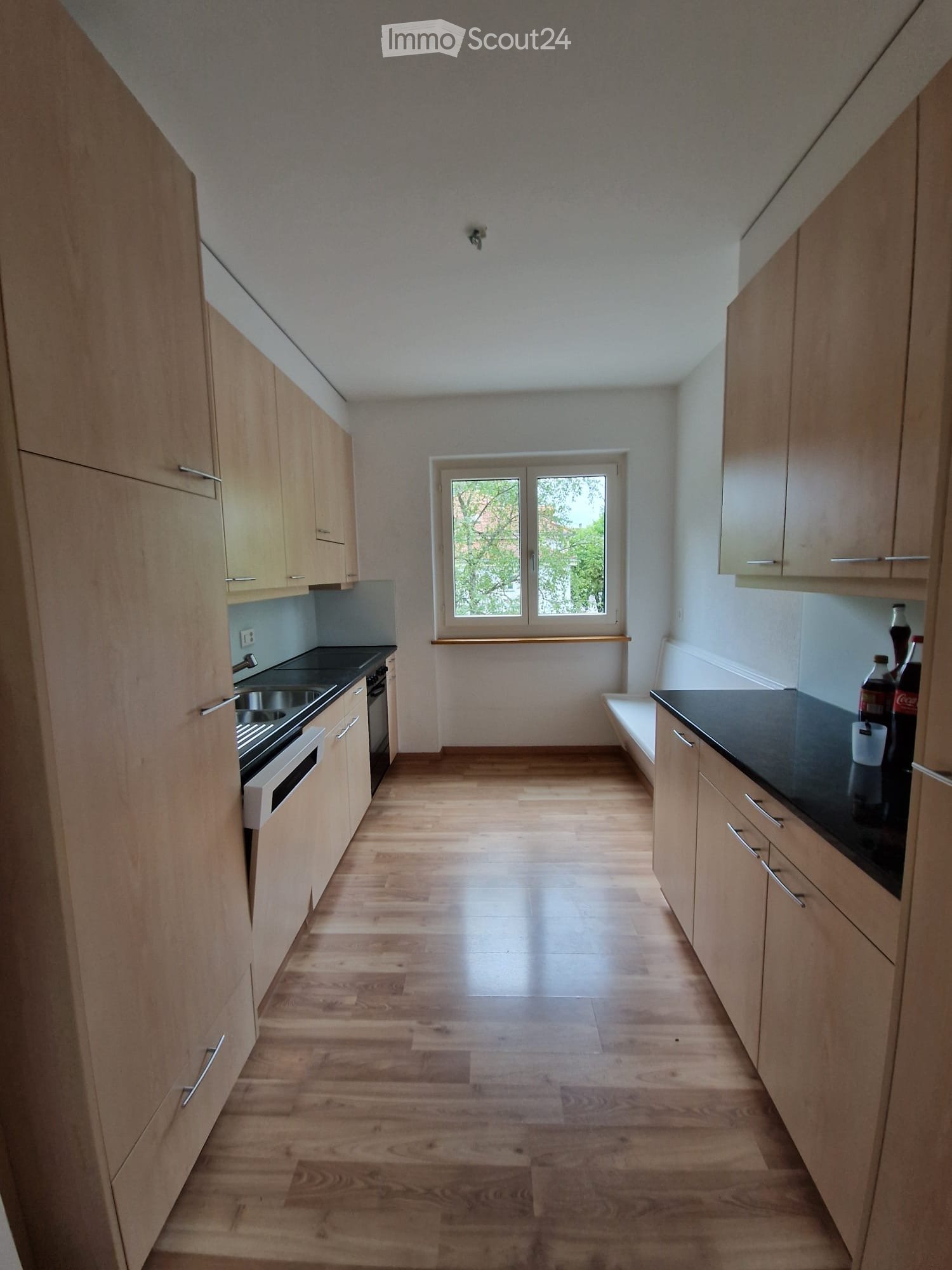 Modern kitchen with light wooden cabinets, black countertop, and a window overlooking greenery.