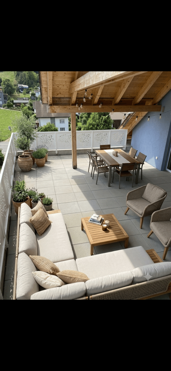 A spacious, well-lit balcony or terrace with a wooden pergola, outdoor seating including a sofa, coffee table, and dining set, surrounded by potted plants and offering a scenic view of a green landscape.