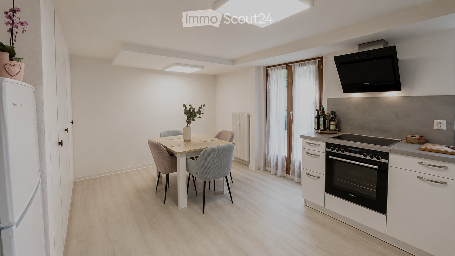 Modern kitchen and dining area with white cabinets, stainless steel appliances, a dining table with four chairs, and a refrigerator. The space features light wood flooring and large windows with white curtains.
