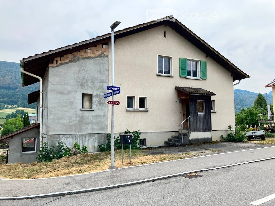 Two-story house with beige walls, green shutters, and a wooden entrance. The roof shows exposed wooden beams on one side. A street sign reading "Rehgasse" is visible in front.