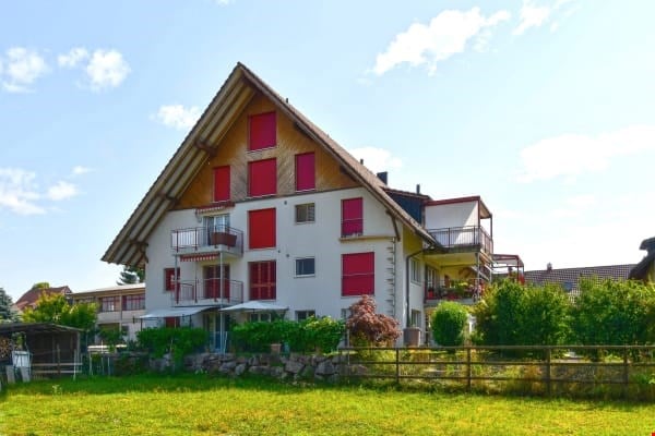 Two-story house with a pitched roof, red shutters, balconies, and a well-maintained garden.