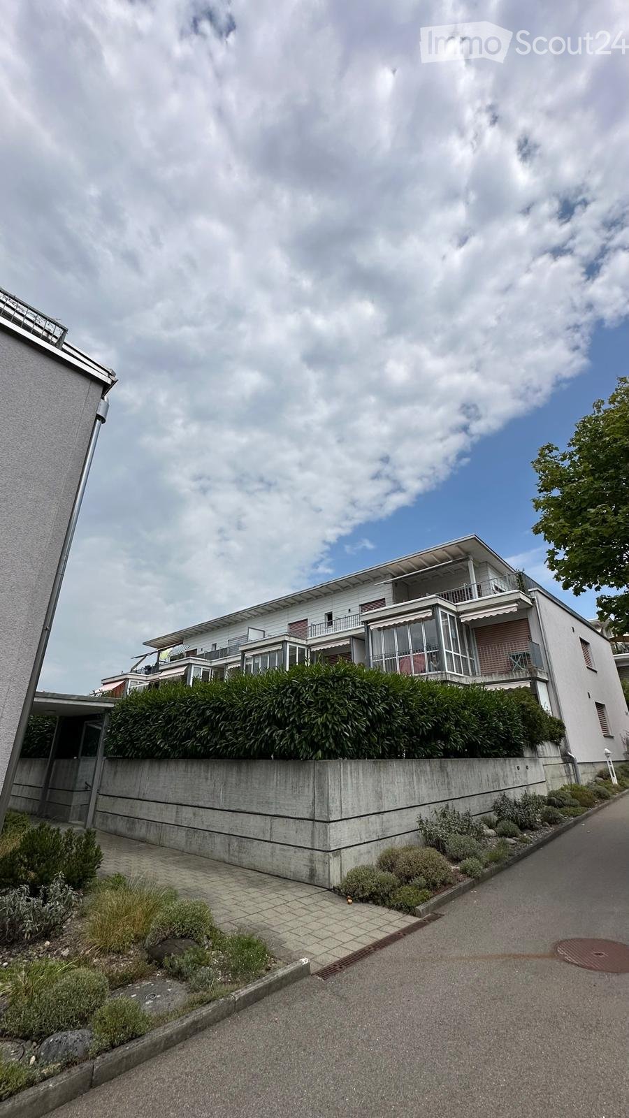 Modern two-story house with white exterior, large windows, balconies, and a well-maintained garden area with shrubs and small plants.