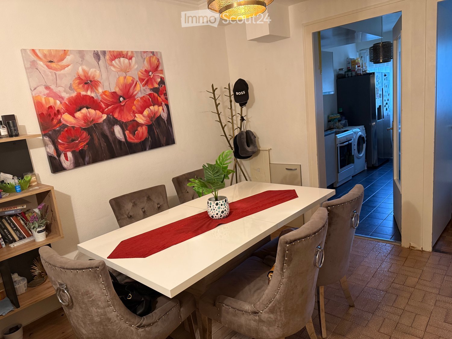 Dining room with a white dining table, six grey chairs, a potted plant, a large painting on the wall, and a shelf with books and decorative items. An open doorway leads to a kitchen area with an oven, refrigerator, and washing machine.