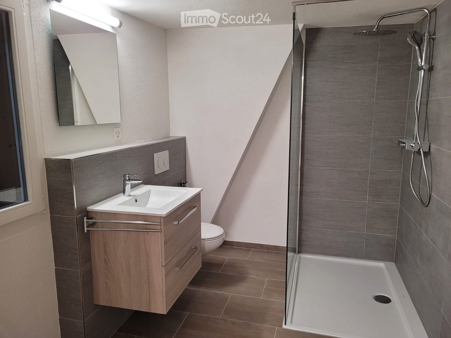 Modern bathroom with a wooden vanity, white sink, chrome faucet, mirror, toilet, and a walk-in shower with a glass enclosure and modern showerhead.
