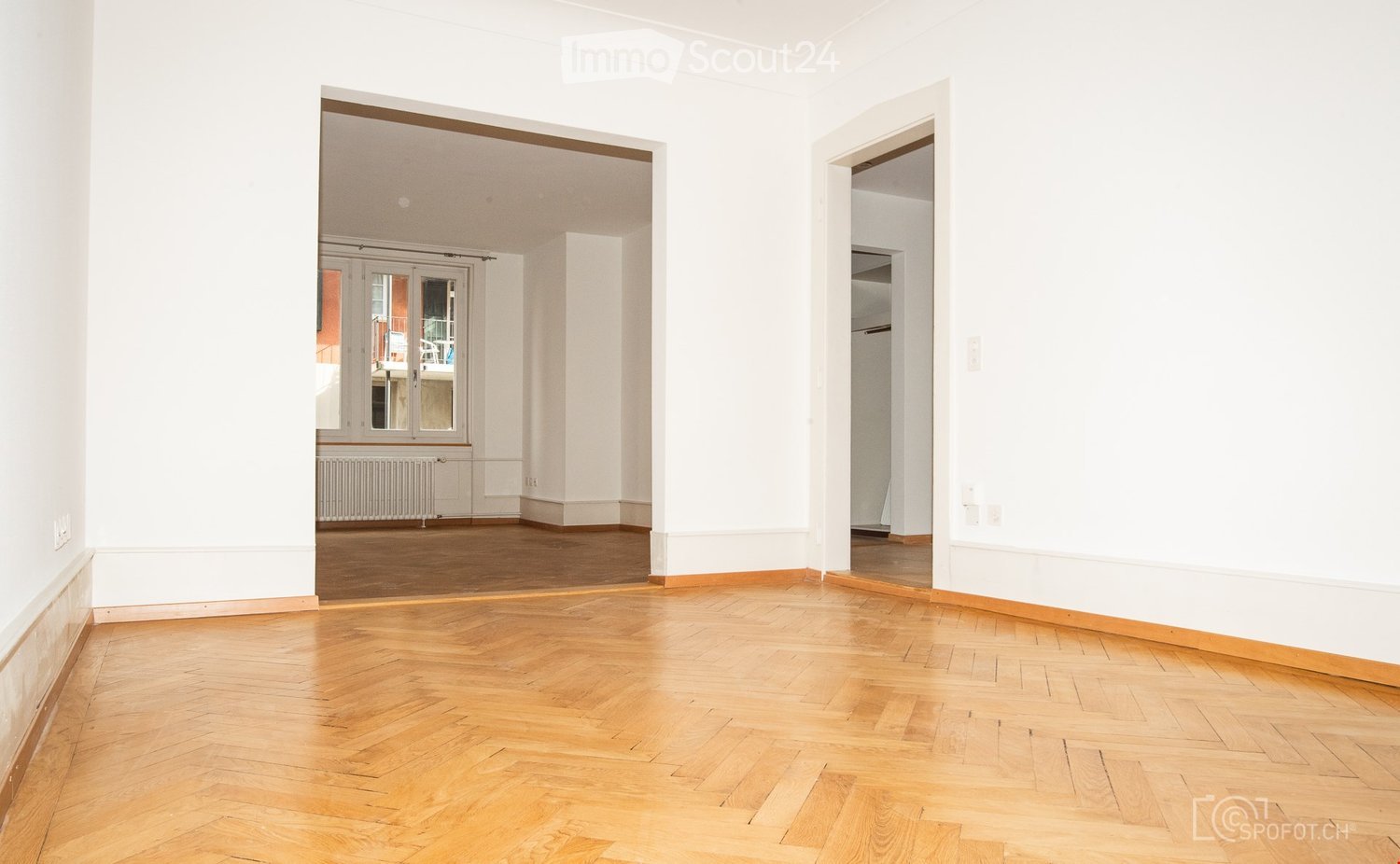 Empty room with light wooden flooring and white walls, featuring an open doorway leading to another room with large windows and a radiator.