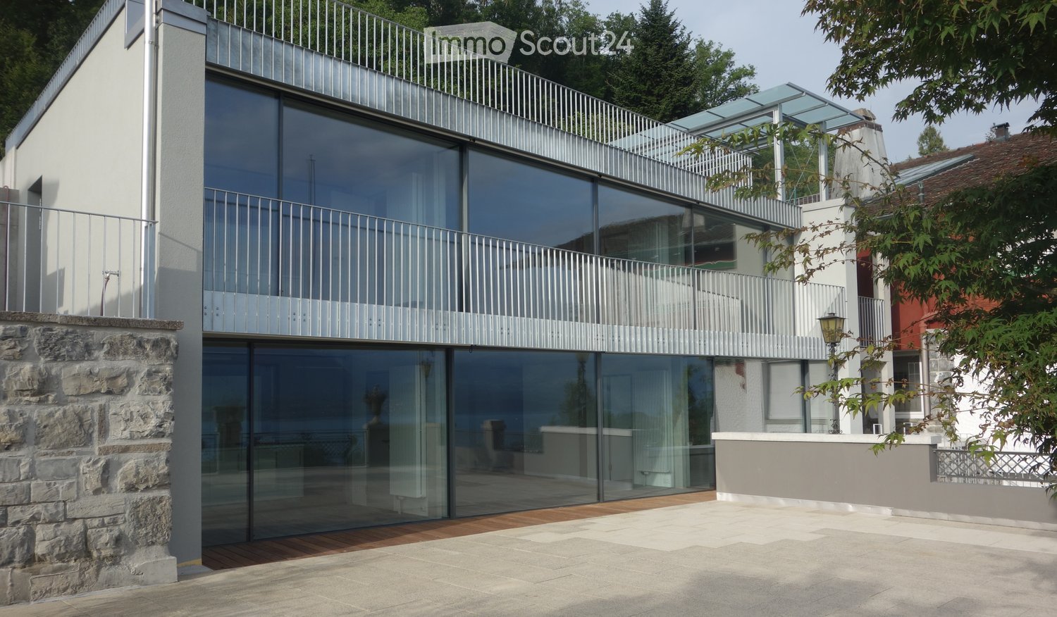 Modern two-story house with large glass windows, metal balconies, and a stone base. The ground floor features a wooden deck area.
