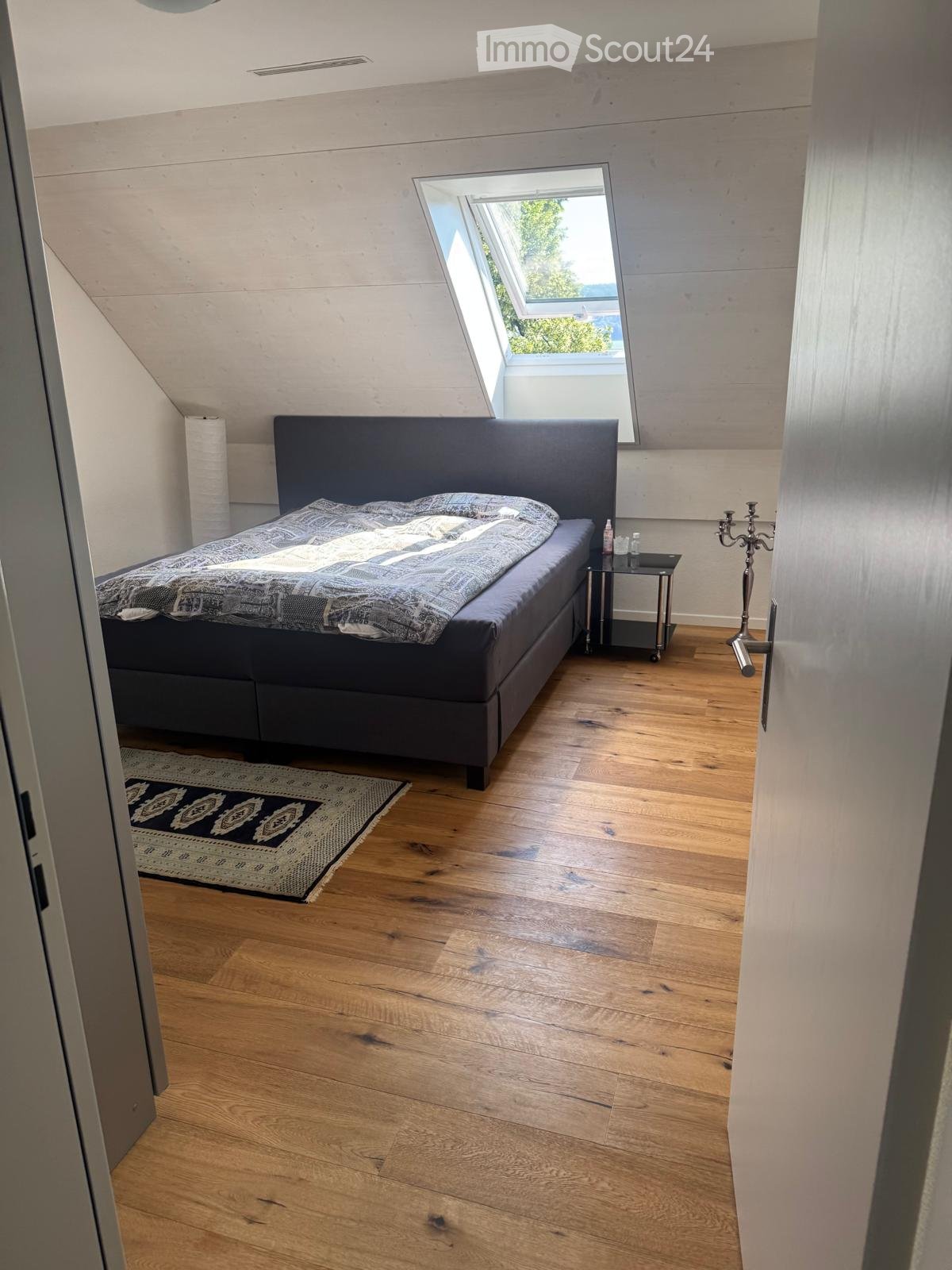 Modern bedroom with wooden flooring, a large bed with dark headboard, nightstand, and a patterned rug.