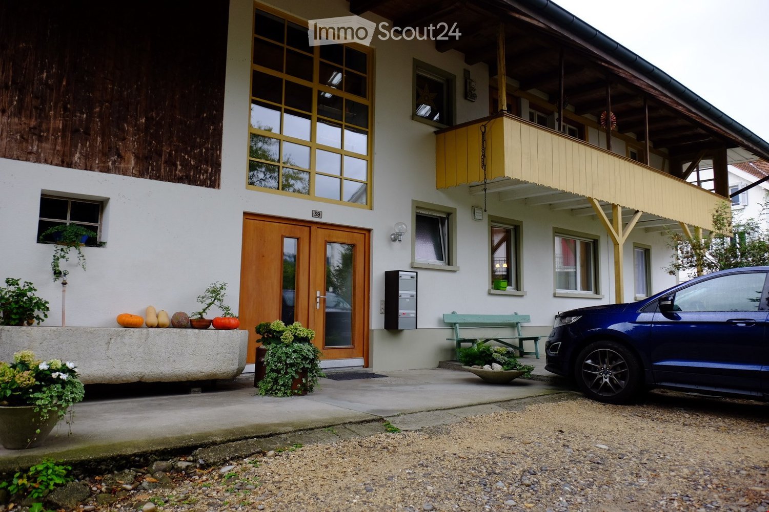 Two-story house with white walls, wooden balcony, large windows, and a blue car parked in front.