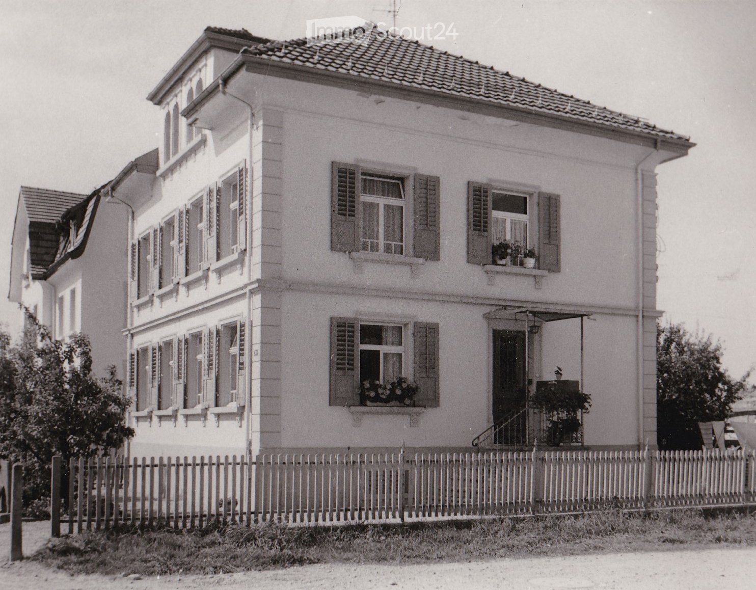Two-story house with white exterior, multiple windows with shutters, small balcony, and a wooden fence in front.