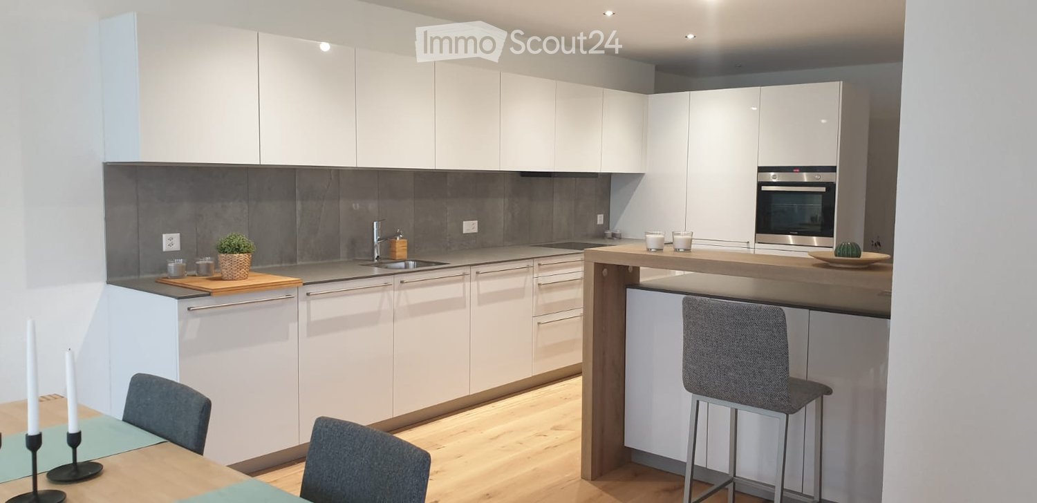 Modern kitchen with white cabinets, grey backsplash, wooden countertops, and integrated appliances. Includes a dining area with a table and chairs, and a bar area with stools.
