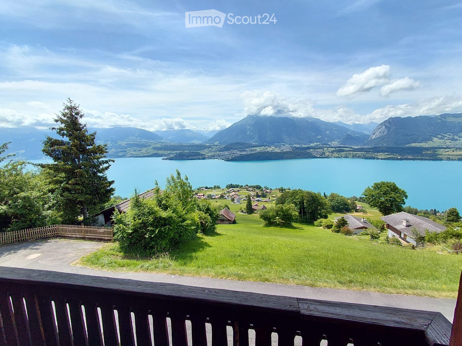 Balcony overlooking a scenic lake and mountain landscape with a wooden railing, green lawn, and distant houses.