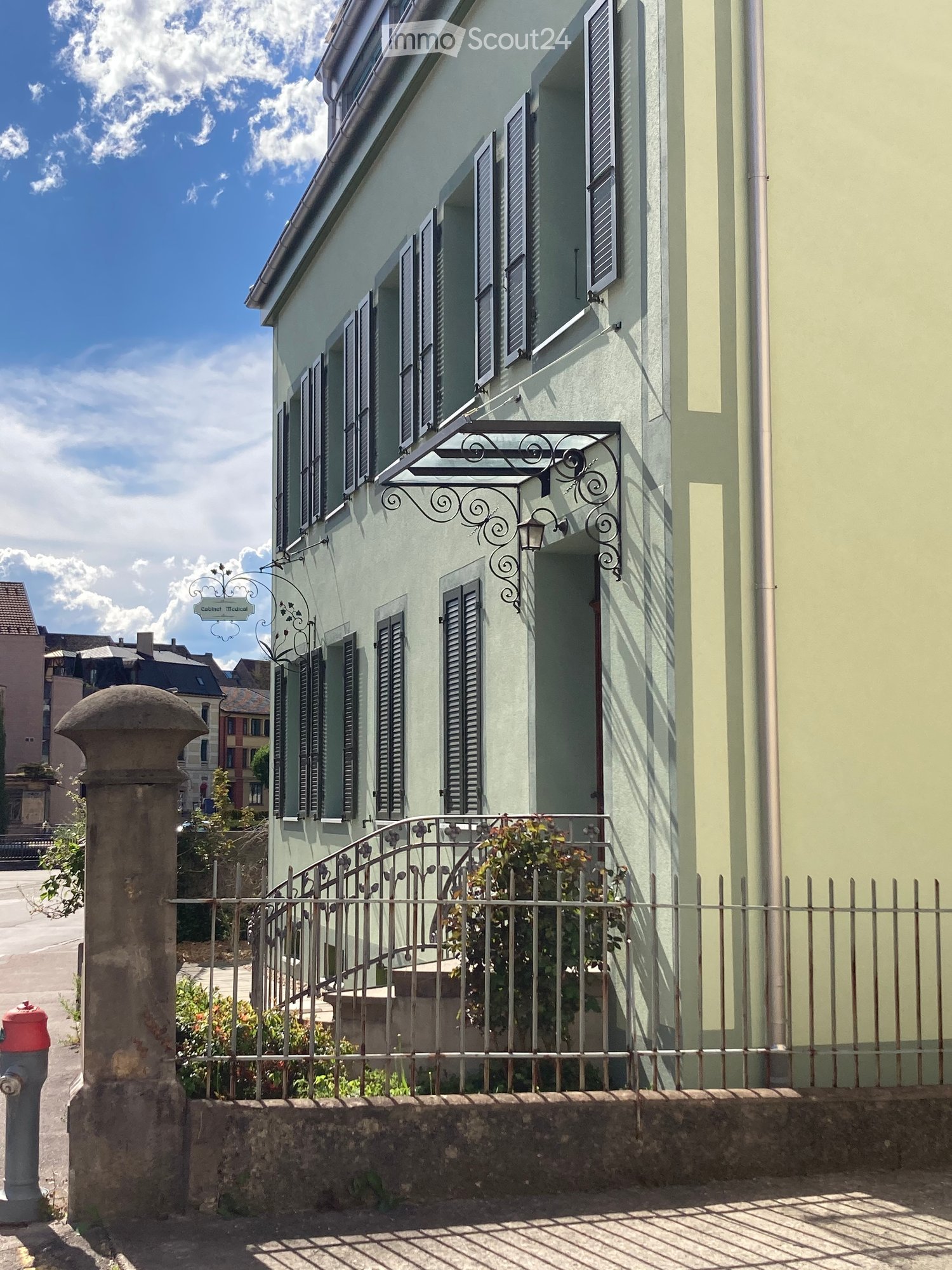 Building. A two-story building with light green walls, multiple windows with shutters, a decorative wrought iron balcony, and a small garden area behind a metal fence.