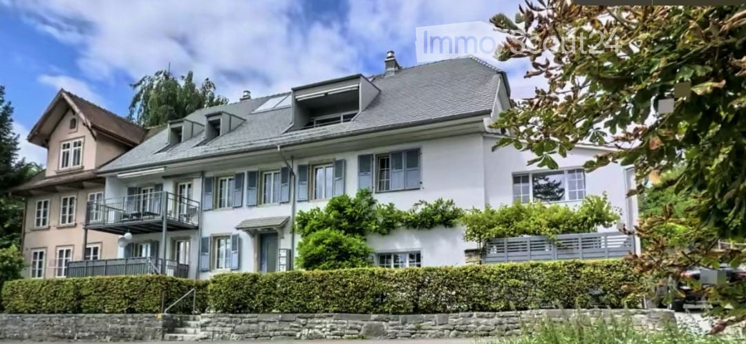 A multi-story residential house with a grey roof, multiple windows, and a balcony on the upper floor. The house is surrounded by greenery and has a stone pathway leading to the entrance.