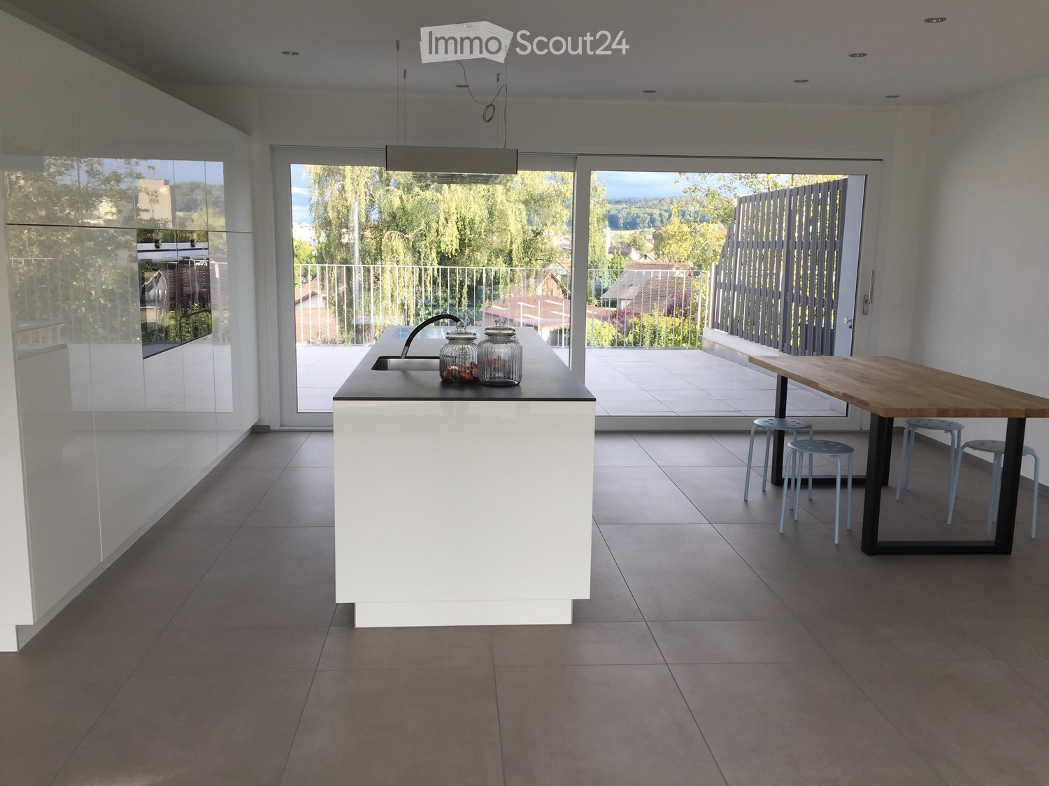Modern kitchen with white island, sink, wooden dining table, and four stools, overlooking a balcony with outdoor view.