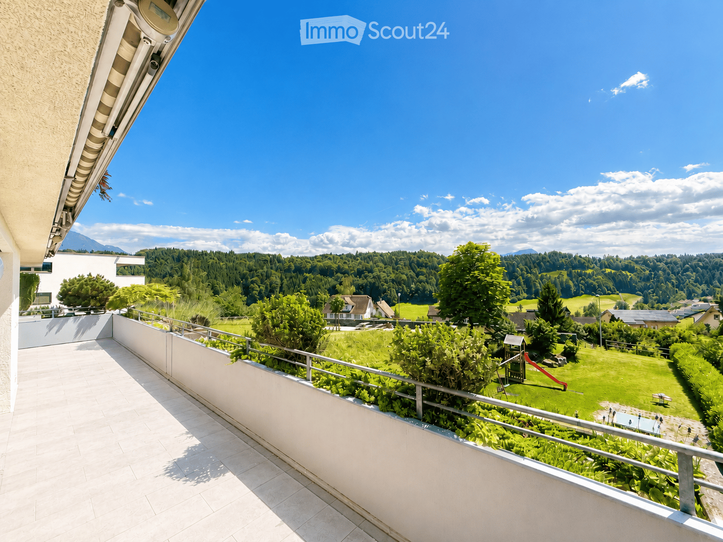 Balcony with tiled floor, metal railing, overlooking a green park area with trees, a playground, and distant mountains.