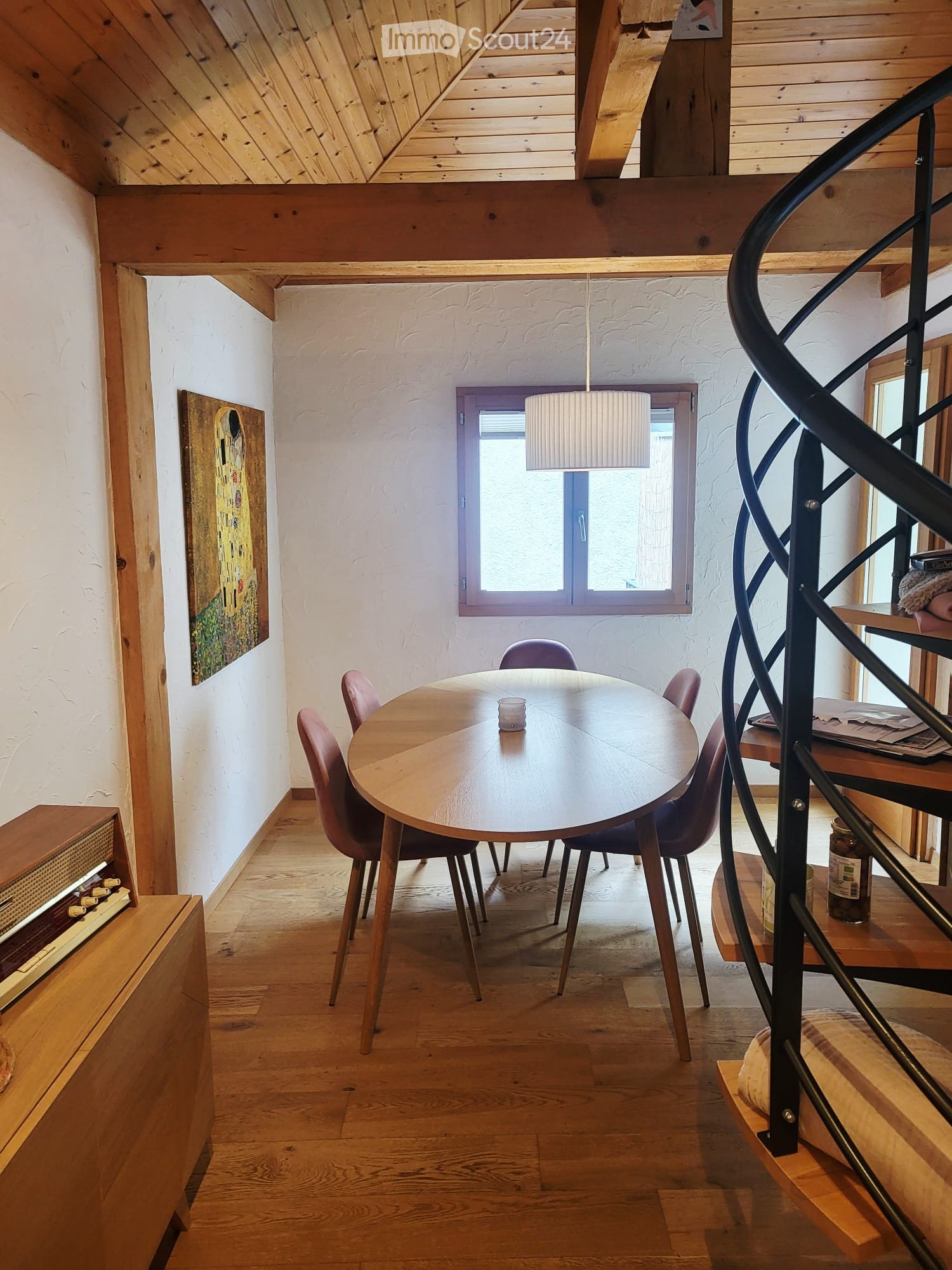 Dining area with a round wooden table, six chairs, a window, a staircase with a black railing, and a painting on the wall.