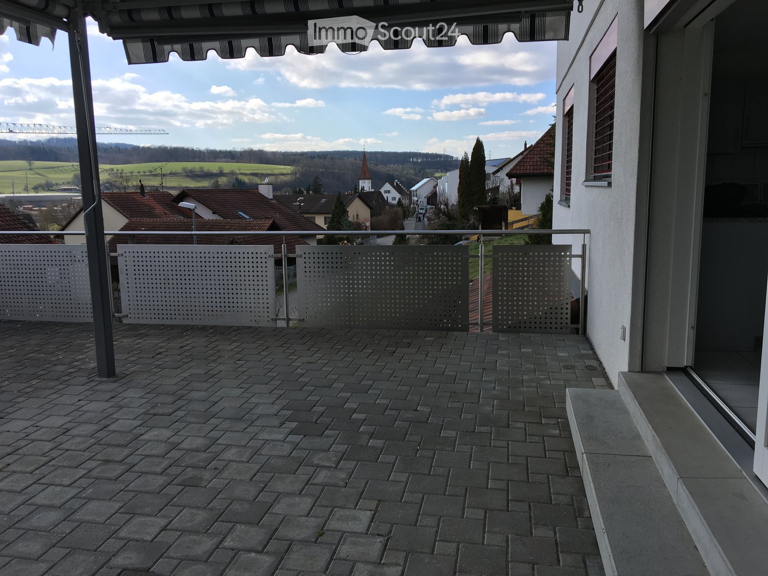 Balcony with brick tiles, metal railing, and a view of a rural landscape with houses and hills.
