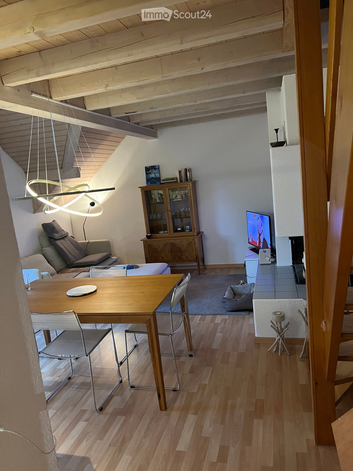 A modern, open-concept living and dining area with exposed wooden beams, a sleek hanging light fixture, a comfortable sofa, a dining table with chairs, a cabinet with glass doors, and a television on a stand.