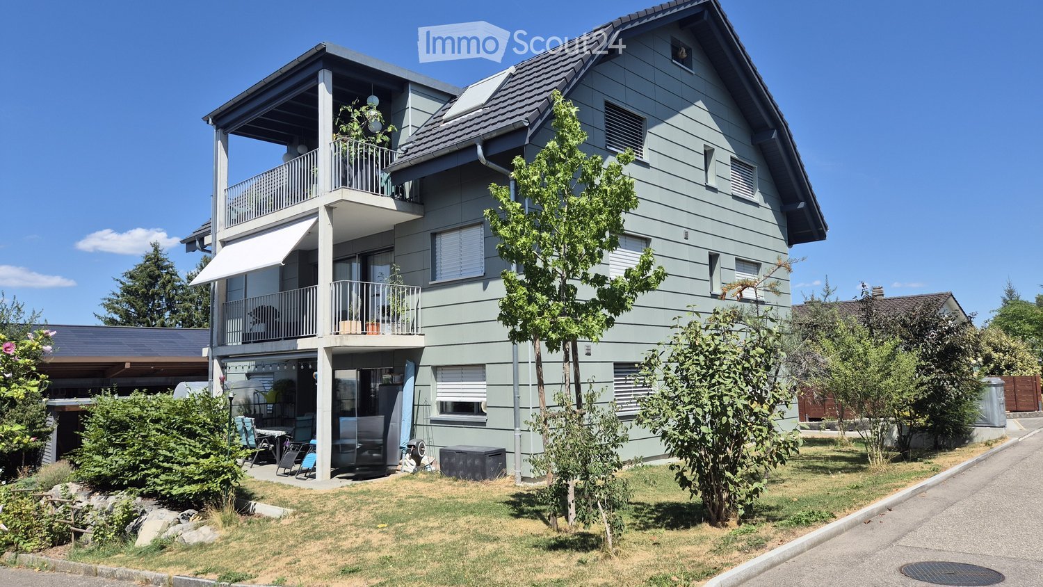 Two-story house with light green siding, multiple windows, balconies, and a small garden area with outdoor furniture.