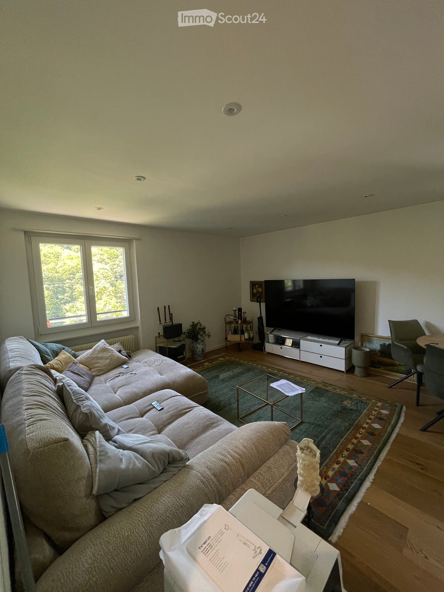 A modern living room with a large sectional sofa, coffee table, TV, and a small table with a plant and candle holder. The room features wooden flooring and large windows letting in natural light.