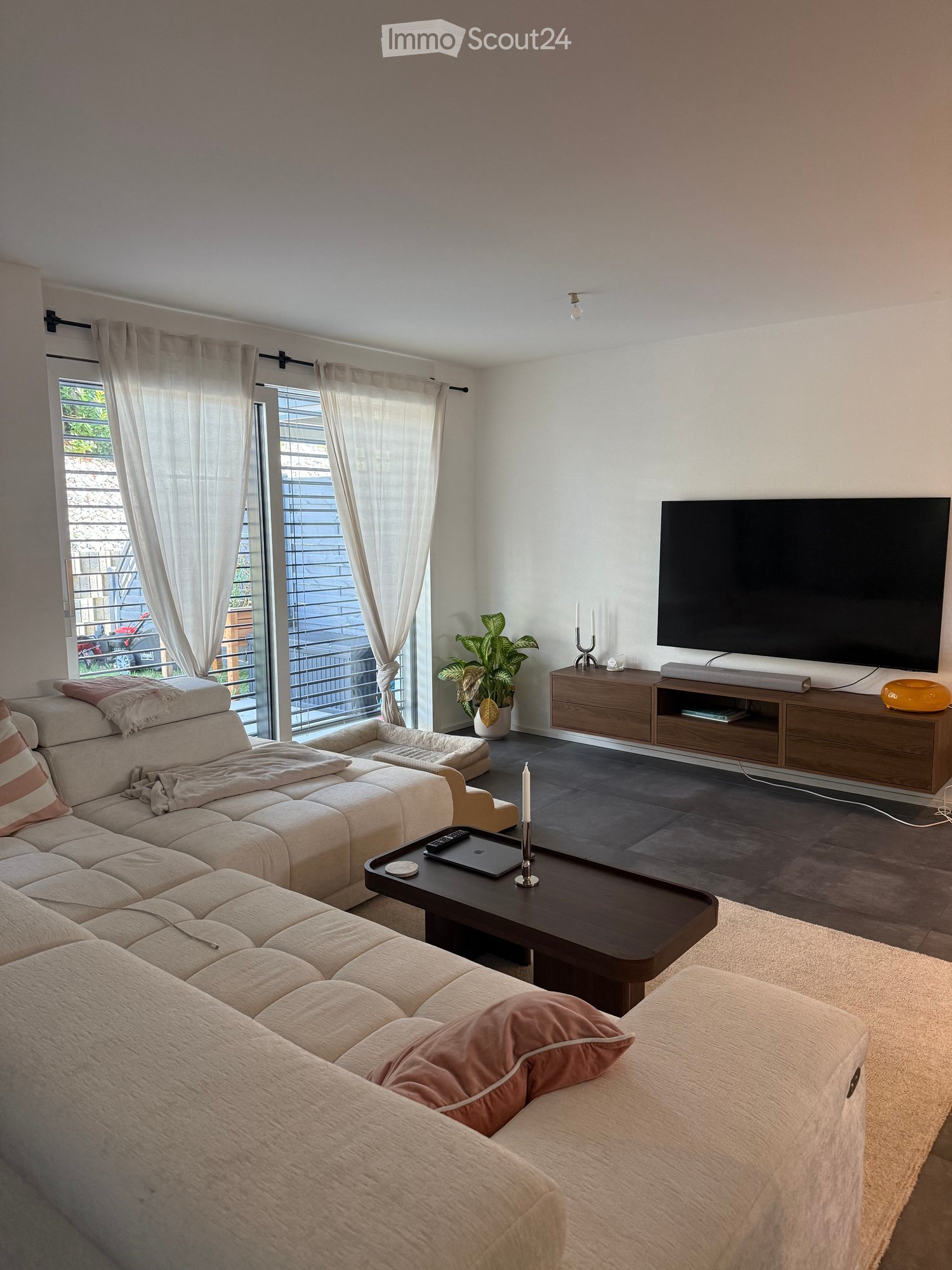 Modern living room with a large sectional sofa, coffee table, flat-screen TV, and a potted plant near the window. The room features light-colored walls, dark flooring, and large windows with sheer curtains.