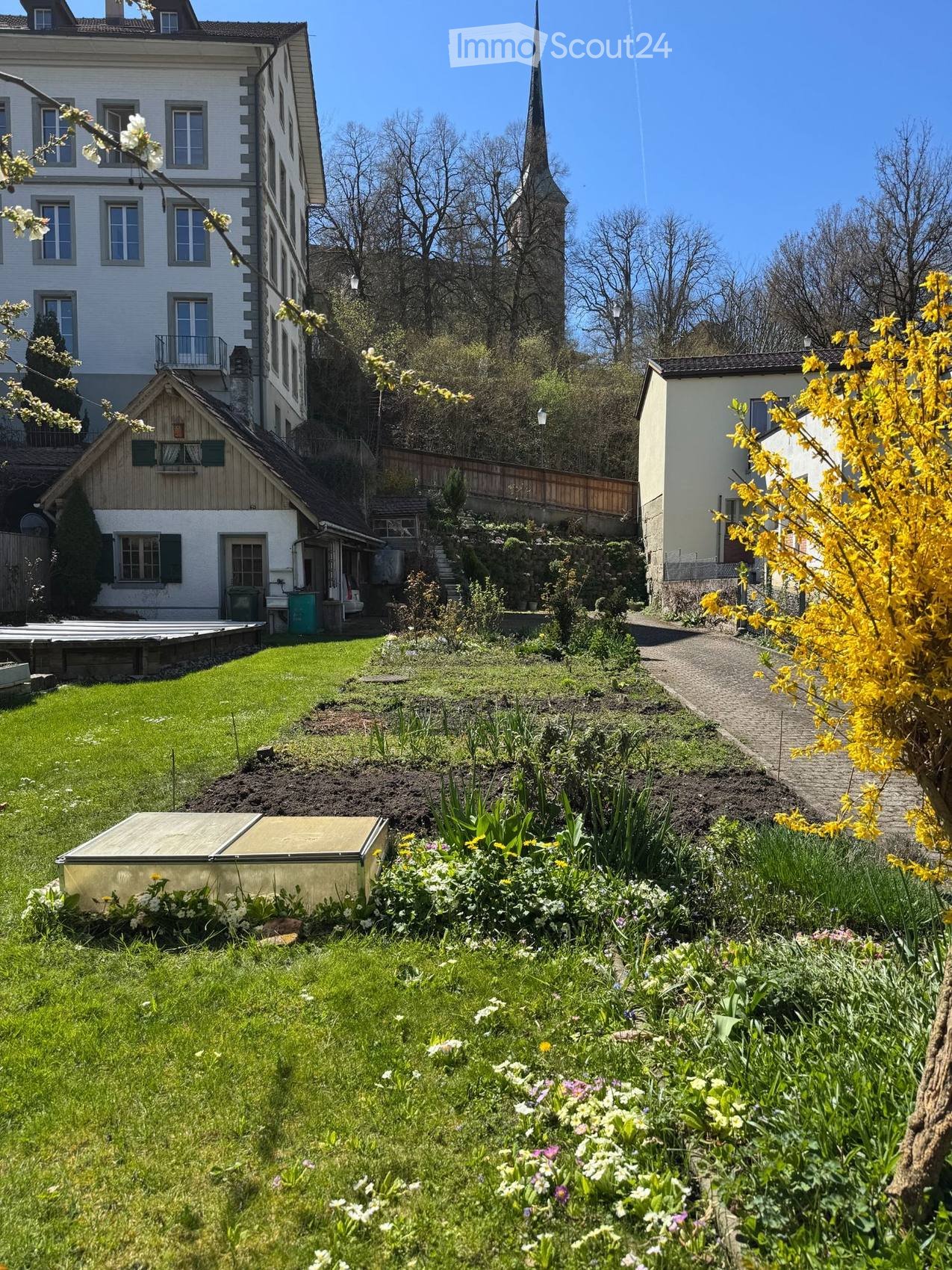 Well-maintained garden with a lawn, flower beds, a gravel path, and surrounding buildings including a tall white house and a smaller wooden structure.