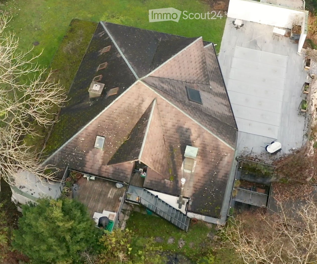 Aerial view of a multi-story house with a complex roof structure, surrounded by greenery and a driveway.