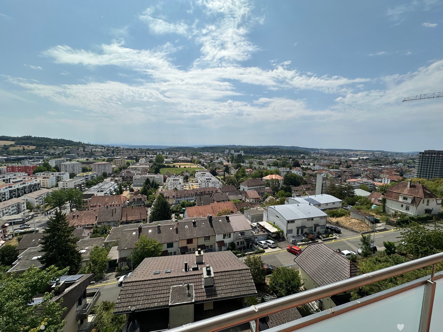 A panoramic view from a balcony or terrace overlooking a suburban neighborhood with houses, trees, and a distant cityscape under a partly cloudy sky.