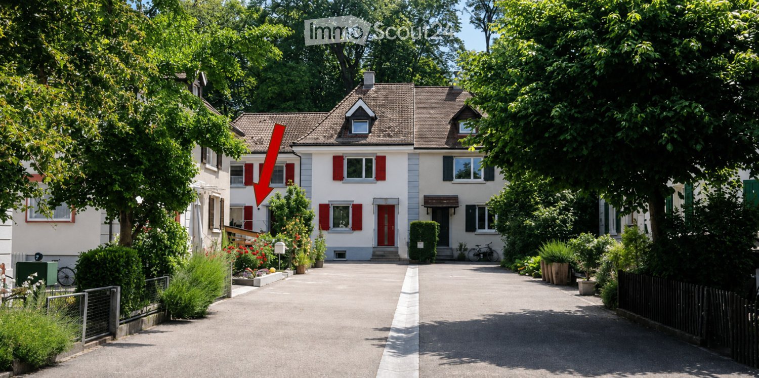 Two-story house with red shutters, white walls, and a tiled roof. The house features a driveway, garden, and is surrounded by trees and greenery.