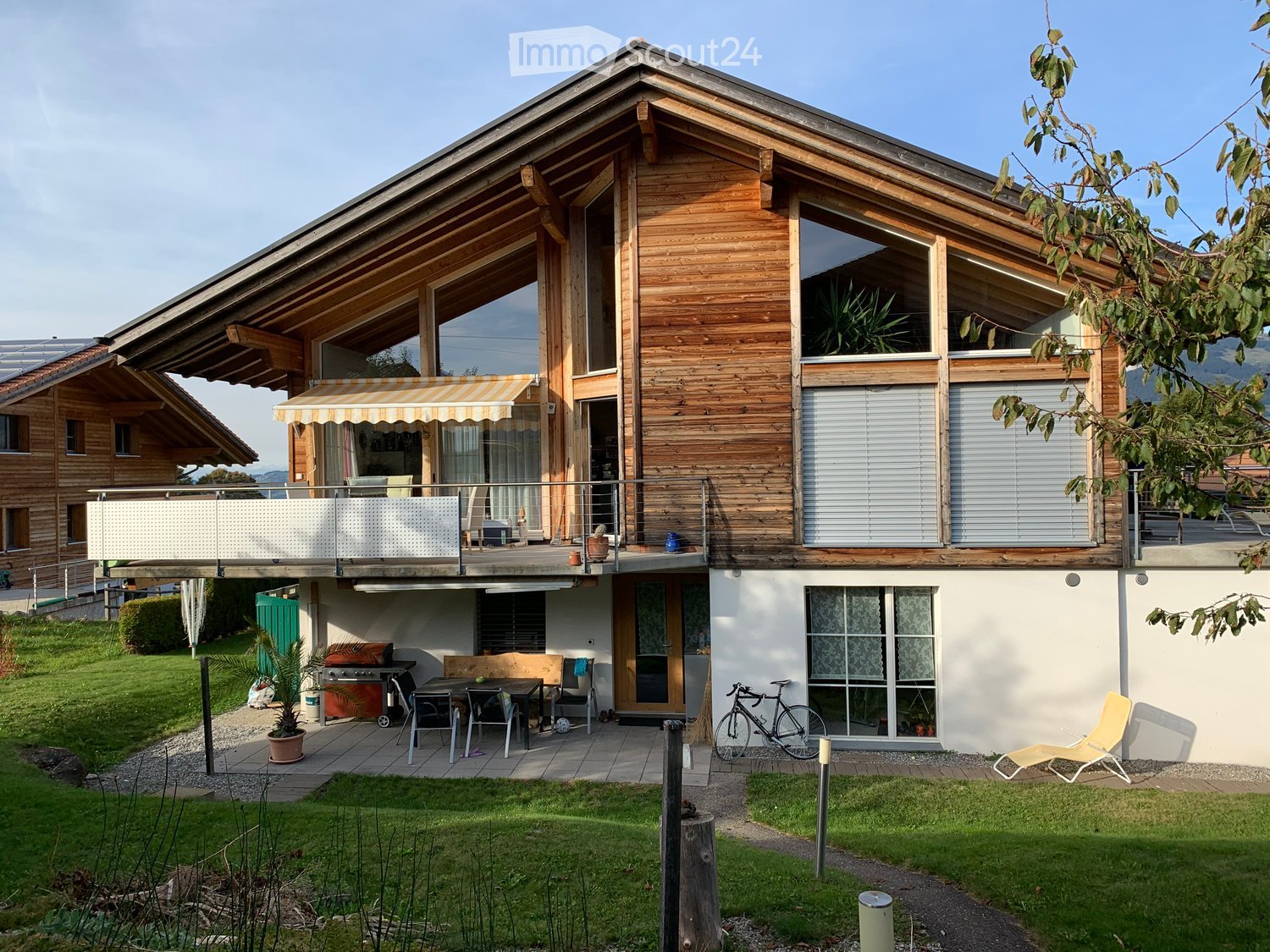 Two-story wooden house with large windows, balcony, outdoor seating area, and garden.