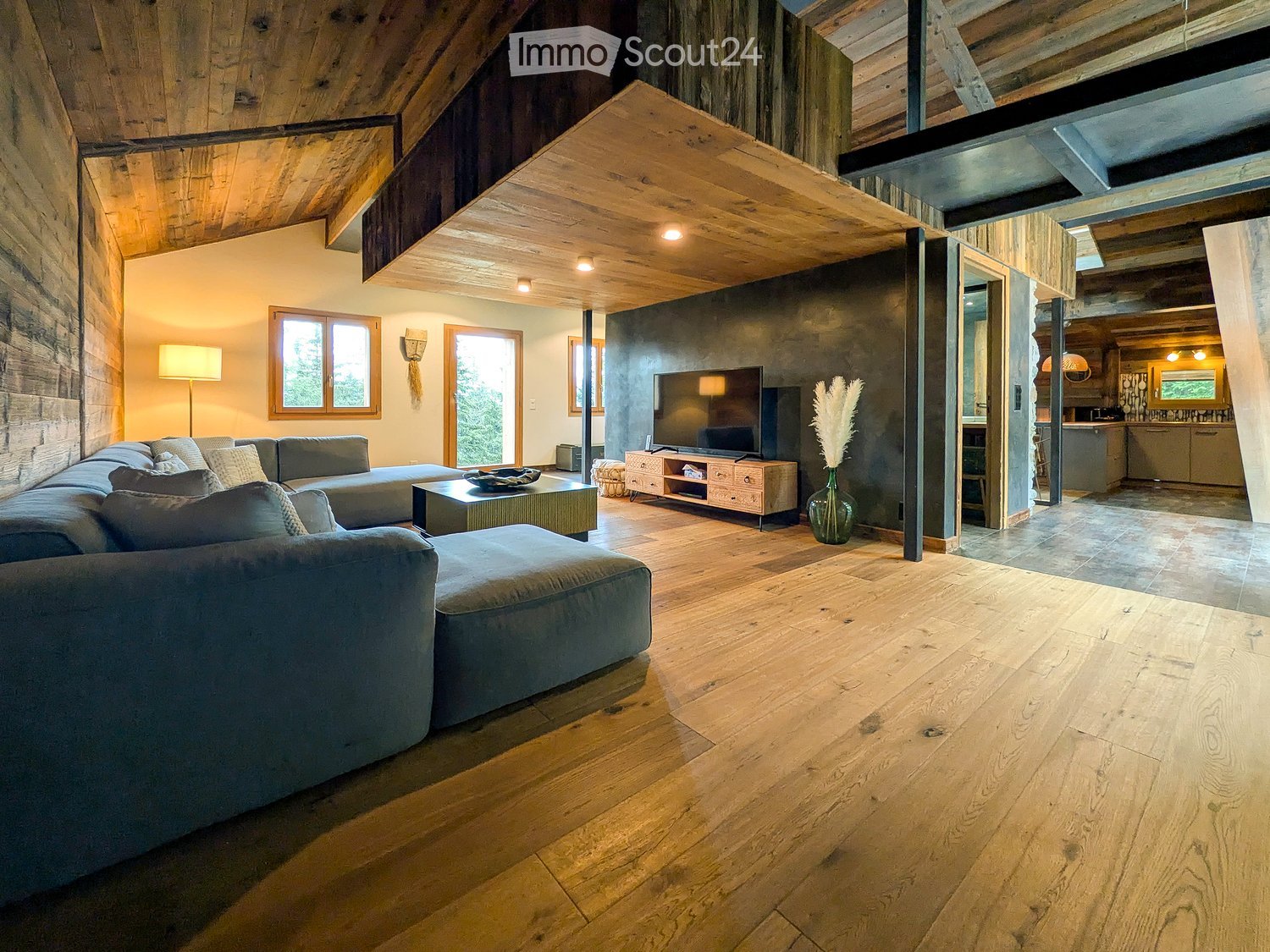 Modern living room with wooden floors, exposed wooden beams, large windows, a sectional sofa, coffee table, TV, and a floor lamp.