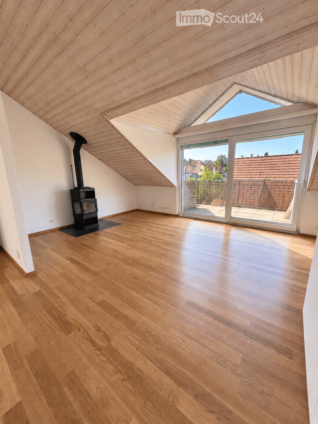 Empty attic room with wooden flooring, white walls, and a skylight window. Features a black wood-burning stove with a chimney pipe and a large sliding glass door leading to a balcony.