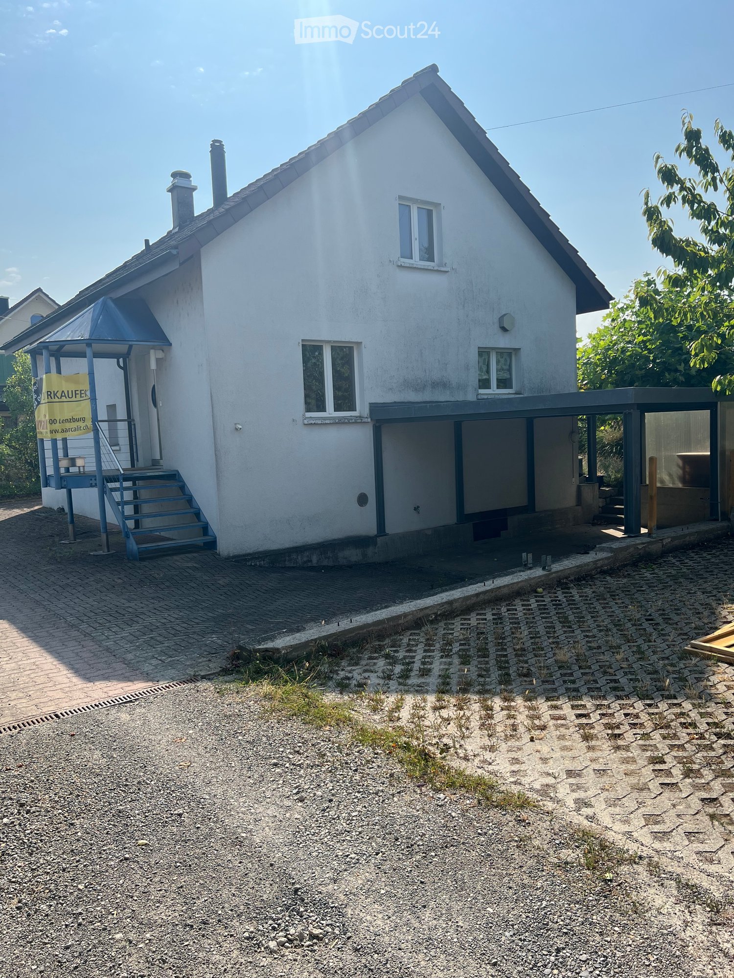 Two-story white house with a blue staircase leading to a covered entrance, multiple windows, and a gravel driveway.