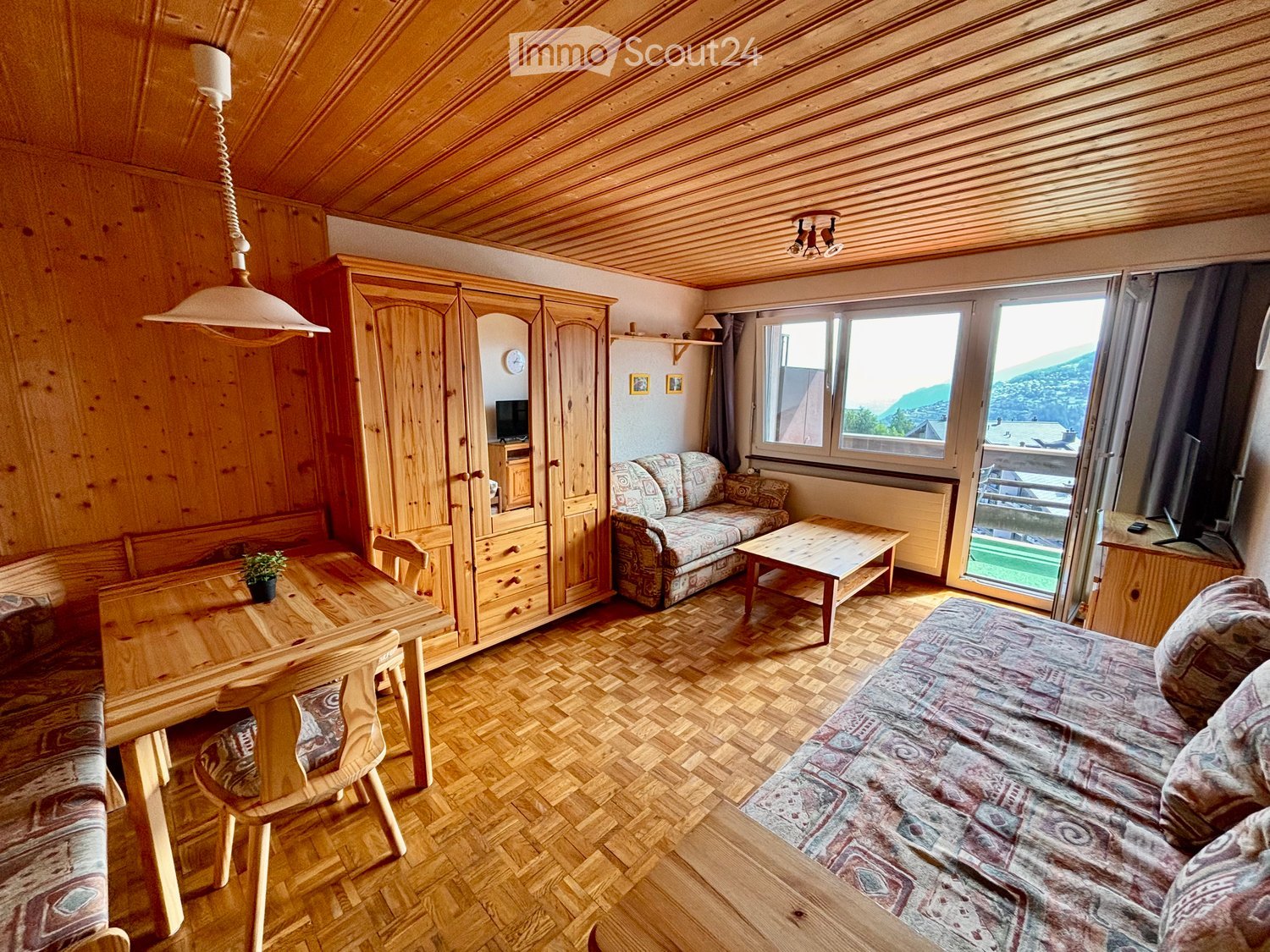 Cozy, wooden-furnished room with a sofa, coffee table, dining table, chairs, wardrobe, and television, overlooking a scenic mountain view.