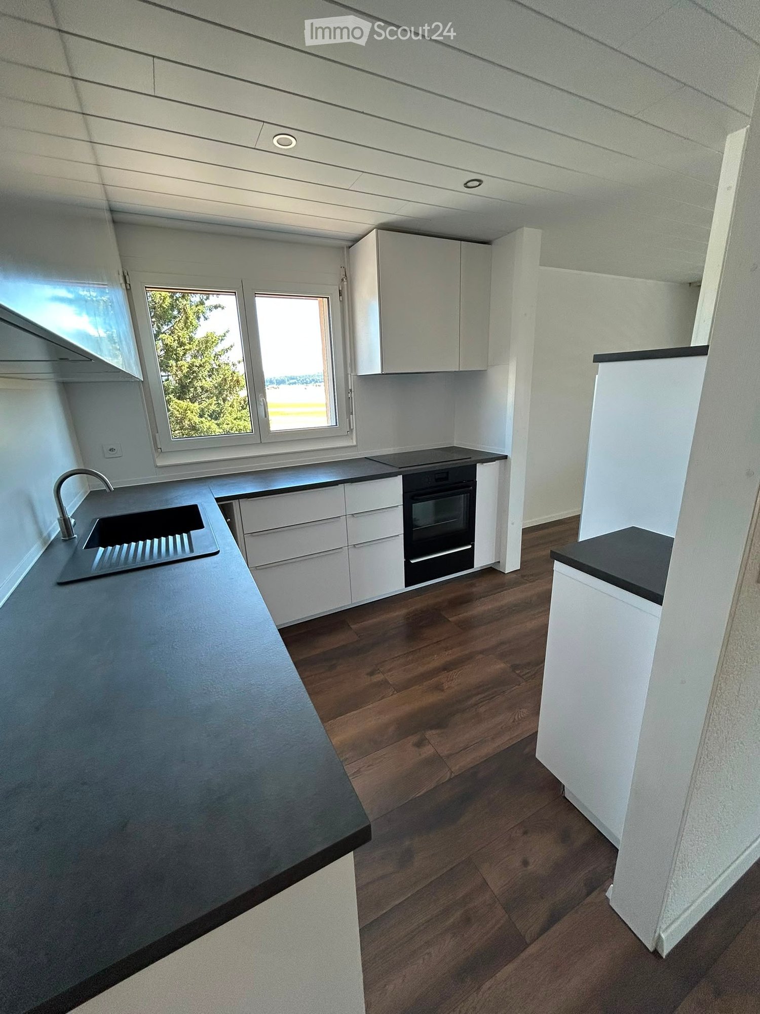 Modern kitchen with white cabinets, dark countertops, wooden flooring, and a large window overlooking greenery.