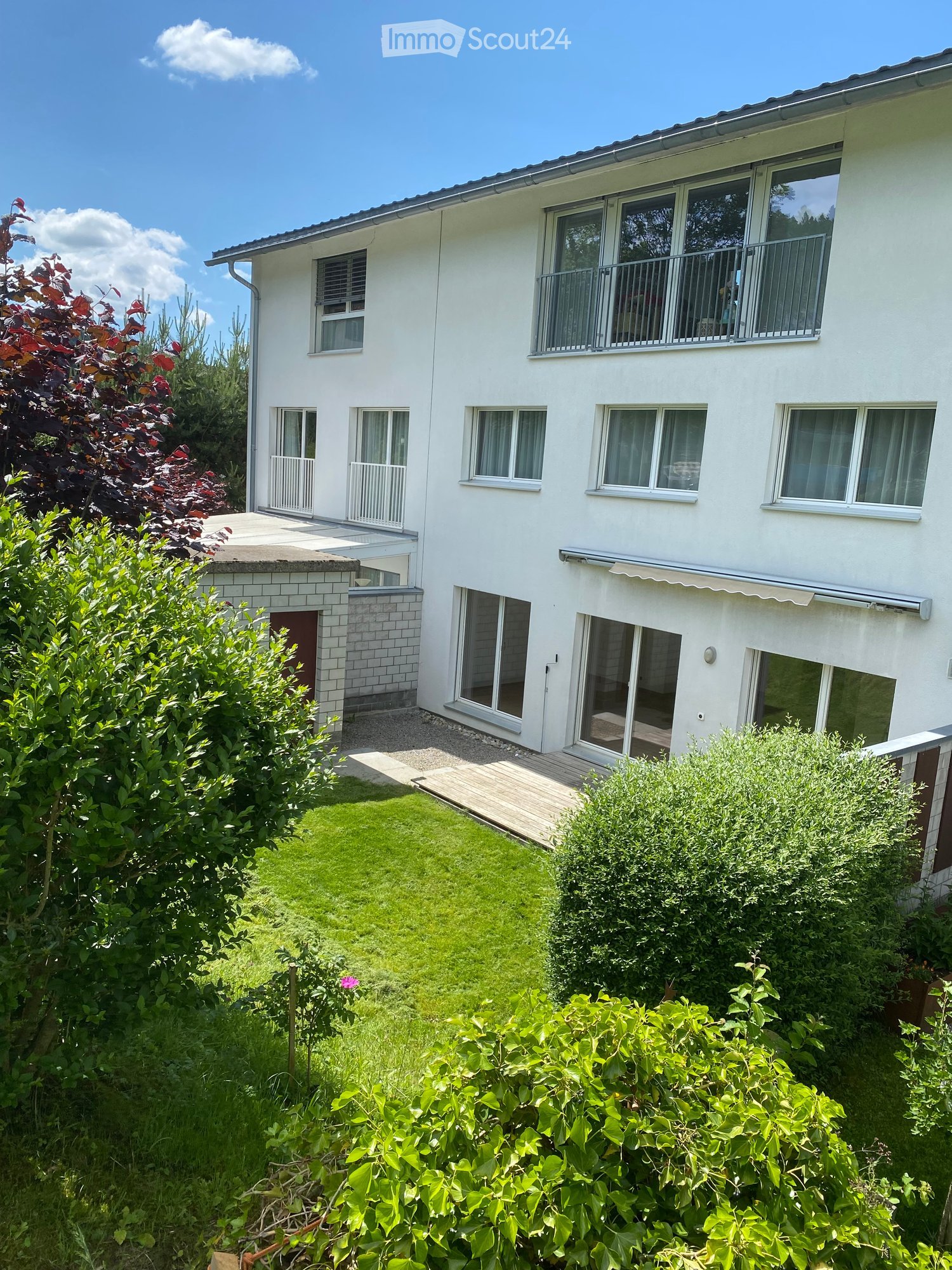 Two-story white house with multiple windows, balconies, and a well-maintained garden with green lawn and various bushes.