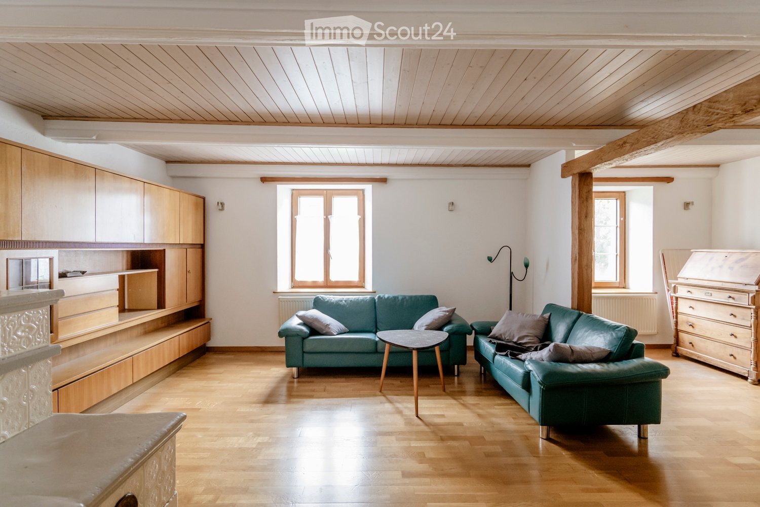 Bright living room with wooden flooring, white walls, and exposed wooden beams on the ceiling. Features two green sofas, a round coffee table, built-in wooden cabinets, and a drawer unit.
