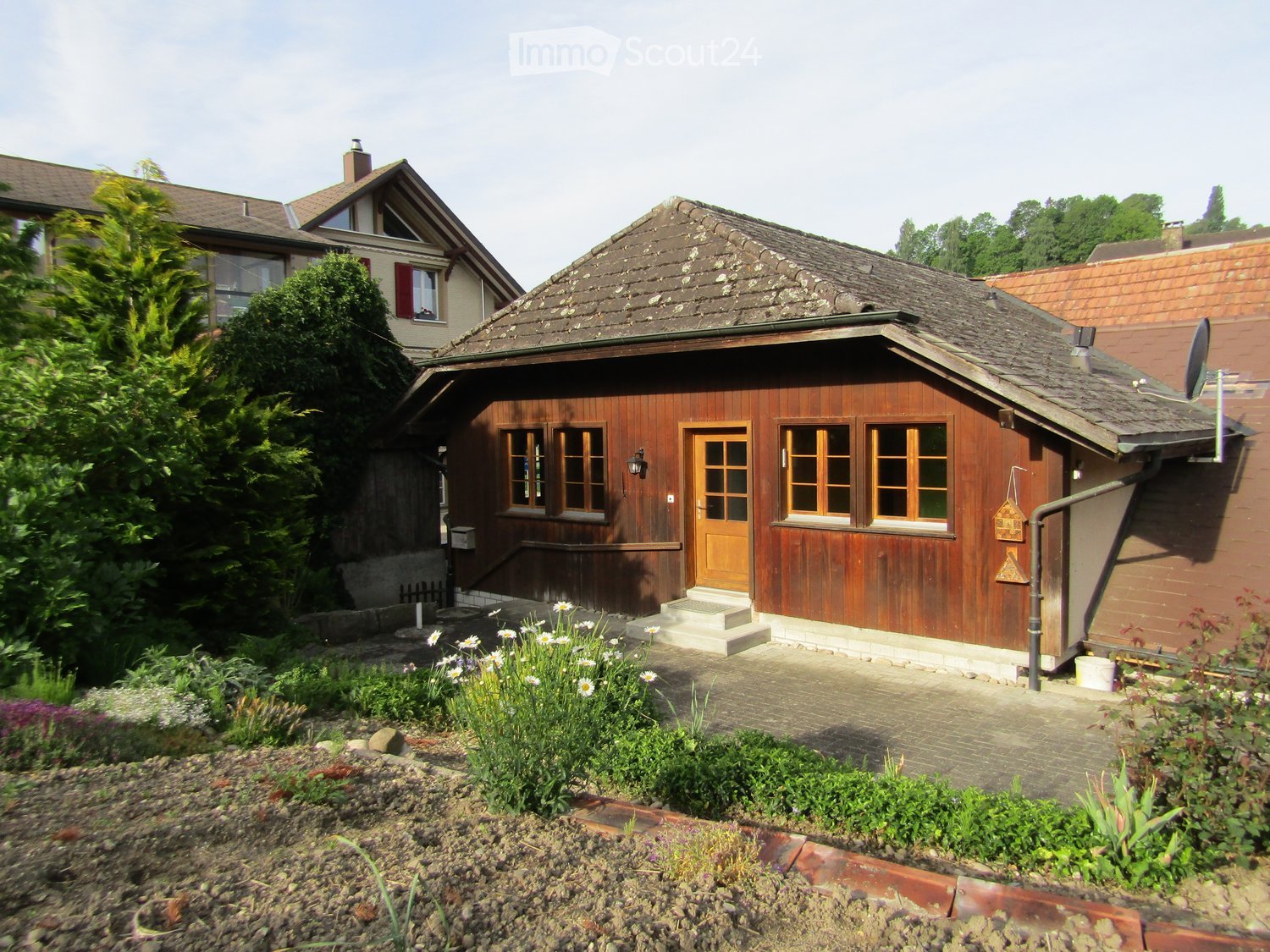 A wooden house with a tiled roof, multiple windows, and a small garden area with flowers and plants.