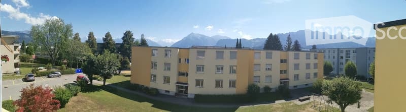 A multi-story residential building with light yellow and cream-colored facades, surrounded by trees and parked cars, with a scenic mountain backdrop.