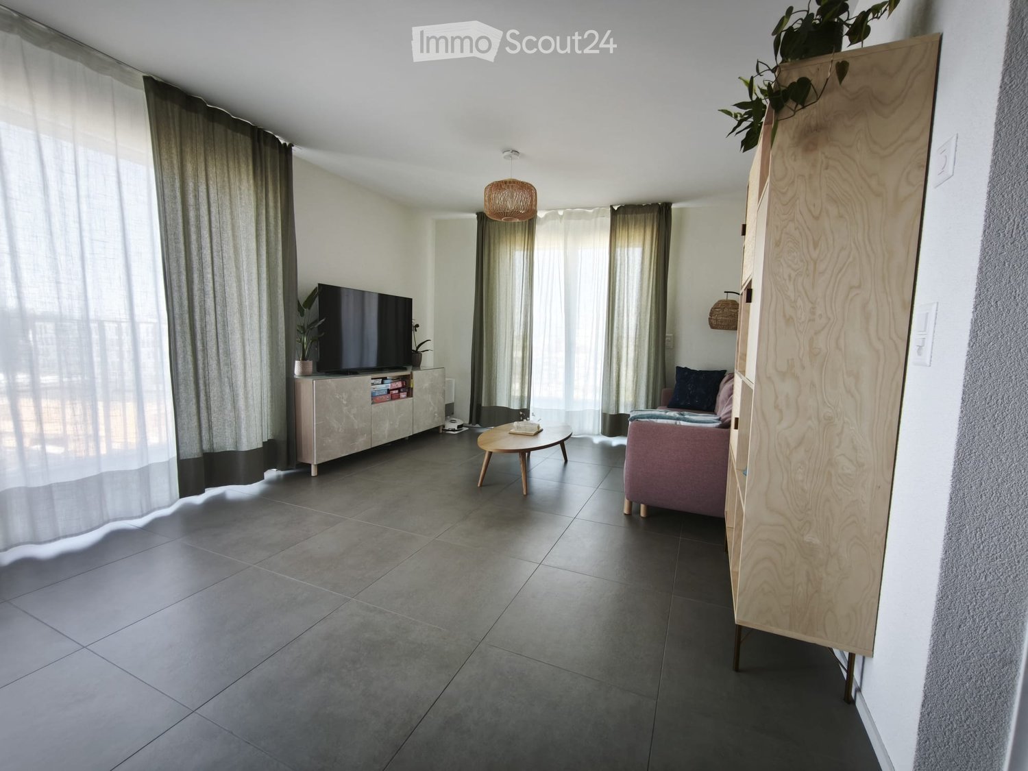 Modern living room with grey tiled flooring, large windows with curtains, a pink sofa, a round wooden coffee table, a television on a cabinet, and a floor lamp.