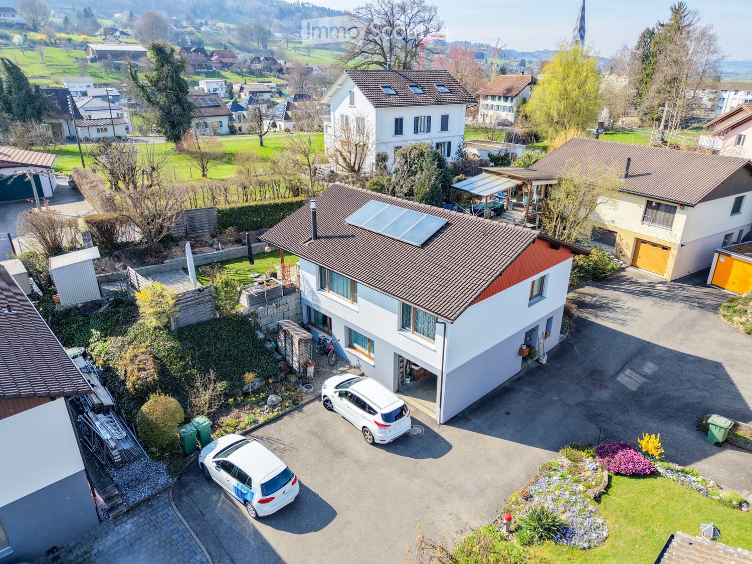 Aerial view of a two-story house with a garage, driveway, and surrounding greenery. Two cars are parked in the driveway, and there are additional houses and greenery in the background.