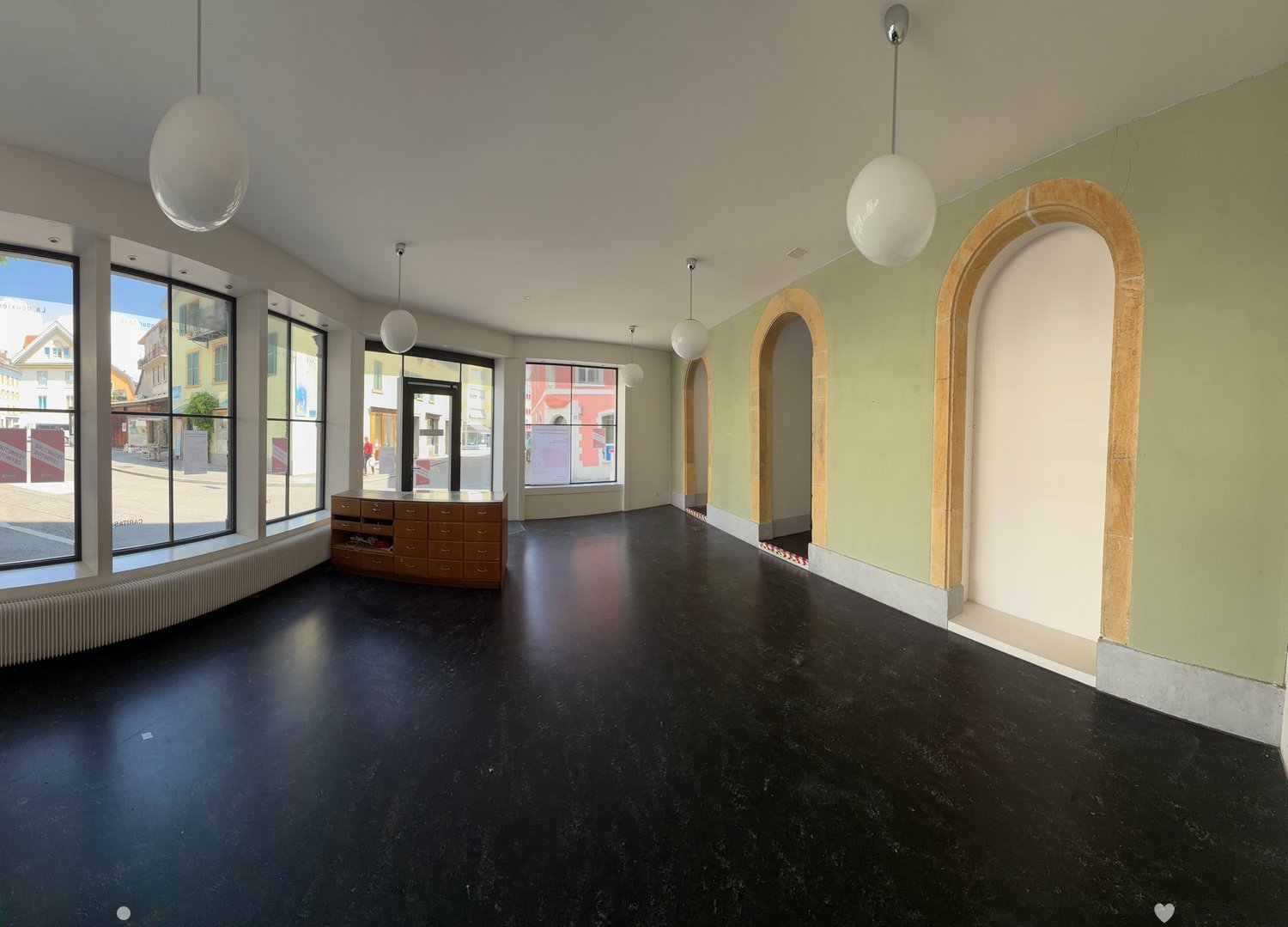 Modern, spacious room with dark flooring, large windows, pendant lights, and a wooden cabinet.