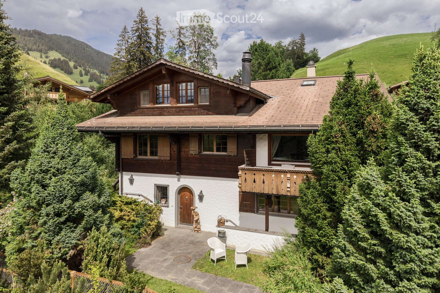 A two-story wooden house with a brown roof and white walls, surrounded by lush greenery and trees, with a small patio area featuring white chairs and a table.